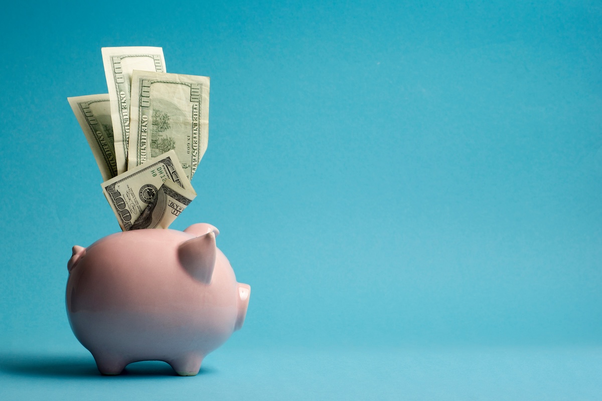 Pink piggy bank with cash sticking out on a blue background, symbolizing saving strategies for building an effective emergency fund.